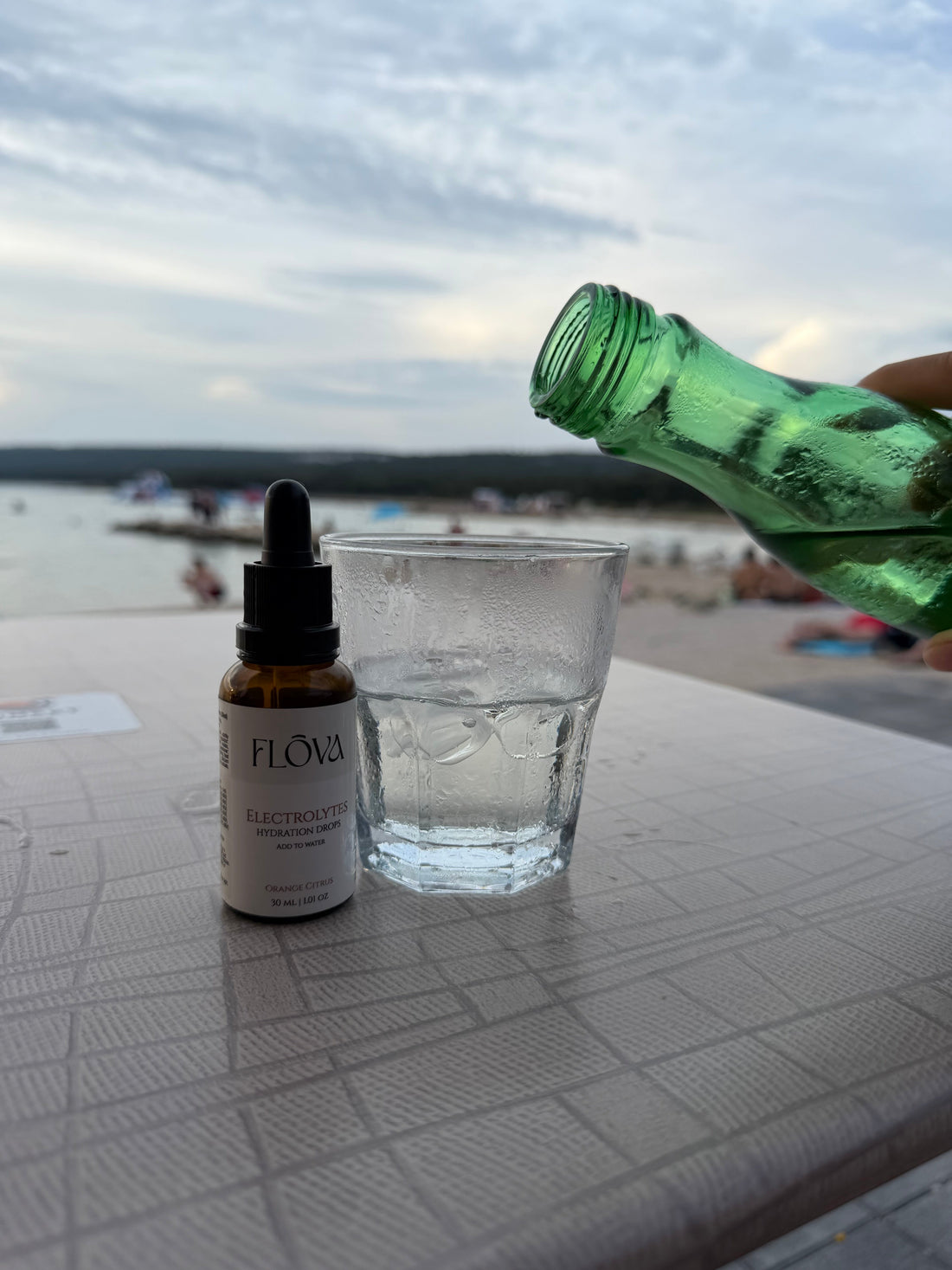 Flōva hydration drops uz čašu vode dok se ulijeva mineralna voda, uz more u pozadini.